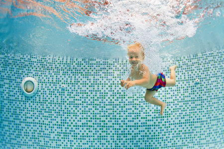 Funny portrait of child learning swimming, dive in blue pool with fun - jumping deep down underwater with splashes. Healthy family lifestyle, kids water sports activity, swimming lesson with parents.の写真素材