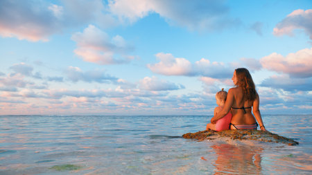 Happy people have fun on summer beach holiday. Young mother with daughter relax at sea water pool. Looking at beautiful view of sunset sky. Healthy family lifestyle, summer travel on tropical island.の写真素材