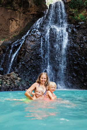 In tropical jungle happy mother with kids stand under waterfall, explore rainforest nature. Travel adventure, hiking activity with child. Lifestyle on family summer vacation and weekend walking tourの写真素材