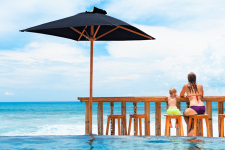 Young mother with happy baby son have fun relaxing at poolside beach bar. Positive girl swim in outdoor infinity pool with ocean view. Healthy travel lifestyle, kids on summer family holiday.の写真素材