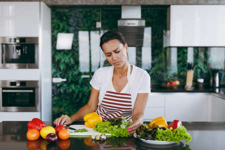 The young confused and thoughtful woman in apron decides what to cook in the kitchen. Healthy food - Vegetable salad. Dieting concept. Healthy lifestyle. Cooking at home. Prepare foodの写真素材