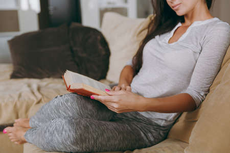 The beautiful young woman at home sitting on the sofa, relaxing in her living room and reading book. Close up.の写真素材