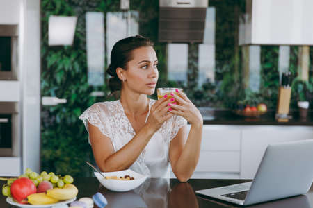 The portrait of beautiful young woman working with laptop while breakfast with cereals and milk and drinking orange juice. Eating at home.の写真素材