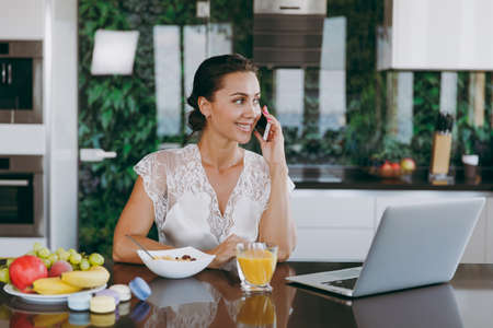 The portrait of beautiful happy woman talking on a mobile phone while breakfast with laptop on the table. Eating at home.の写真素材