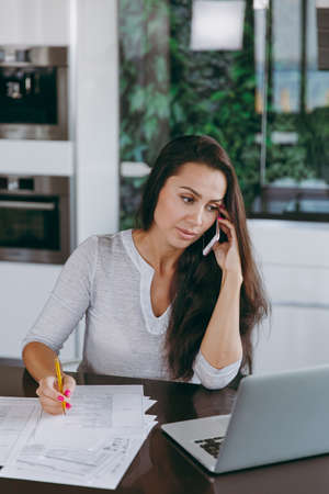 The attractive young modern business woman talking on mobile phone and working with documents and laptop in the kitchen at homeの写真素材