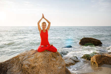 The woman in a red suit practicing yoga on stone at sunrise near the seaの写真素材