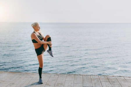 The young beautiful athletic girl with long blond hair in black clothes doing stretching at sunrise over the seaの写真素材