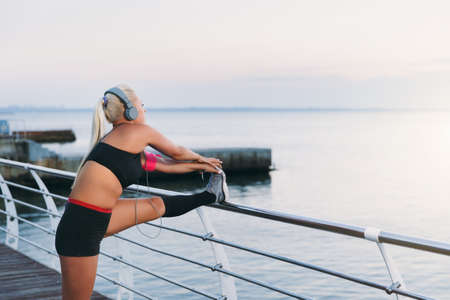 The young beautiful athletic girl with long blond hair in headphones listening to music and doing stretching at sunrise over the seaの写真素材