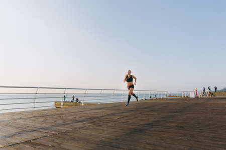 The young beautiful athletic girl with long blond hair in black clothes running at sunrise over the...