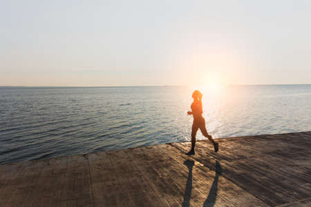 The silhouette of a young beautiful athletic girl with long blond hair in headphones, who listens to music and runs at dawn over the seaの写真素材