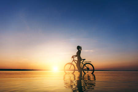 The silhouette of a sporty girl in a suit that sits on a bicycle in the water at sunset on a warm summer day. Fitness concept. Sky backgroundの写真素材
