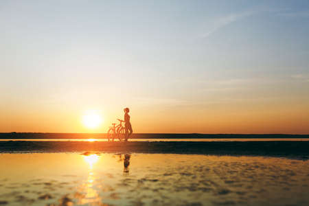 The silhouette of a sporty girl in a suit standing near a bicycle in the water at sunset on a warm summer day. Fitness concept. Sky backgroundの写真素材