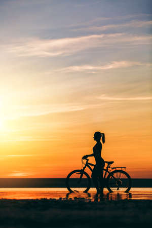 The silhouette of a sporty girl in a suit that sits on a bicycle in the water at sunset on a warm summer day. Fitness concept. Sky backgroundの写真素材