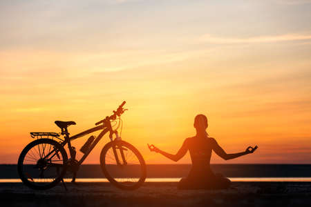 The sports woman doing yoga at sunrise on the sea beach against the background of orange sky and bicycle. Fitness concept. Lotus postureの写真素材