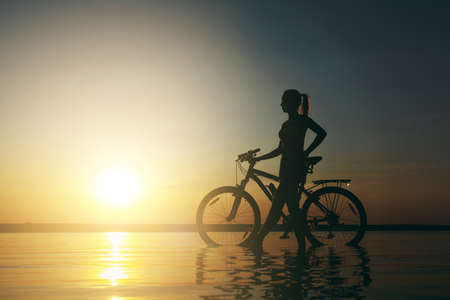The strong blonde woman in a colorful suit stands near the bicycle in the water at sunset on a warm summer day. Fitness concept. Sky backgroundの写真素材