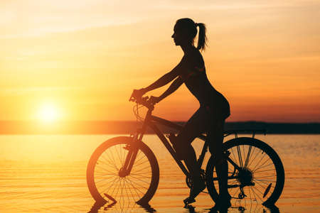 The silhouette of a sporty girl in a suit that sits on a bicycle in the water at sunset on a warm summer day. Fitness concept. Sky backgroundの写真素材