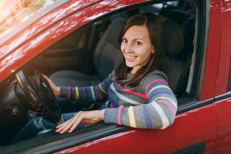 A beautiful young happy smiling European brown-haired woman with healthy clean skin dressed in a striped t-shirt sits in her red car with black interior. Traveling and driving concept.の写真素材