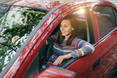 A beautiful young happy smiling European brown-haired woman with healthy clean skin dressed in a striped t-shirt sits in her red car with black interior. Traveling and driving concept.の写真素材