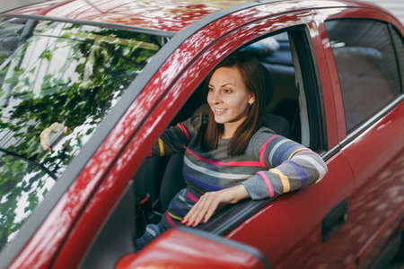 A beautiful young happy smiling European brown-haired woman with healthy clean skin dressed in a striped t-shirt sits in her red car with black interior. Traveling and driving concept.の写真素材