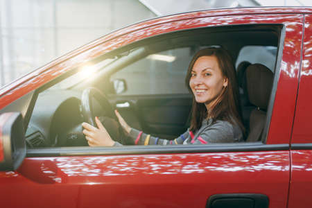 A beautiful young happy smiling European brown-haired woman with healthy clean skin dressed in a striped t-shirt sits in her red car with black interior. Traveling and driving concept.の写真素材