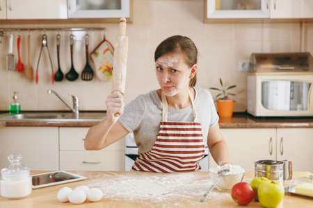 The young perplexed and confused woman sitting at a table with rolling pin and flour and going to prepare a Christmas cakes in the kitchen. Cooking home. Prepare food.の写真素材