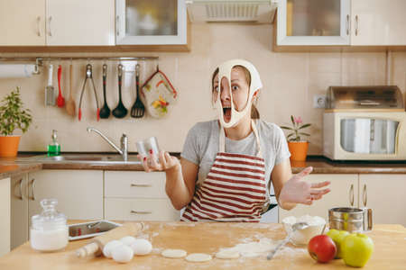 The young funny cheerful and smiling woman puts on a dough with holes on her face and has fun in the kitchen. Cooking home. Prepare food.の写真素材