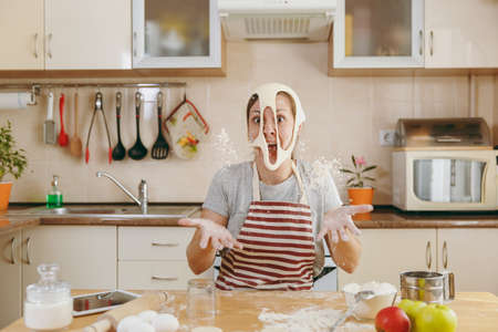 The young funny cheerful and smiling woman puts on a dough with holes on her face and has fun in the kitchen. Cooking home. Prepare food.の写真素材