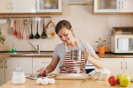 The young beautiful happy woman sifts flour with a sieve and looking for a recipe for Christmas cakes in tablet in the kitchen. Cooking home. Prepare food.の写真素材