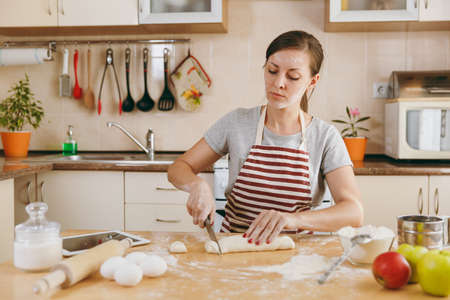 The young beautiful happy woman sitting at a table, cuts a dough with a knife into pieces and going...