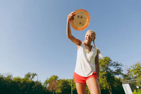 Blonde attractive sporty woman dressed in white T-shirt and orange shorts throwing flying disk to small funny dog, which catching it on the green grass outdoors in park on sky backgroundの写真素材