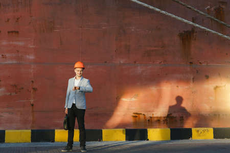 The handsome young unshaven successful business man in gray suit and protective construction orange helmet holding case, looking time on watch, standing in sea port against cargo rusty ship backgroundの写真素材