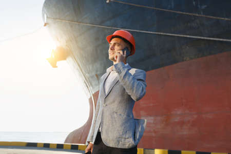 Handsome unshaven successful business man in gray suit and protective construction orange helmet holding tablet, talking on mobile phone, standing in sea port against cargo rusty ship backgroundの写真素材