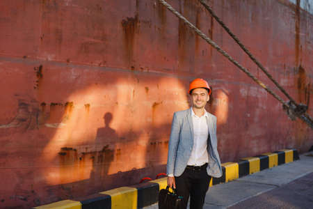 Sunny morning portrait of handsome young unshaven successful business man in gray suit and protective construction orange helmet holding case, standing in sea port against cargo rusty ship background.の写真素材