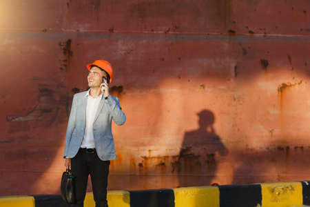 Handsome young unshaven successful business man in gray suit and protective construction orange helmet holding case, talking on mobile phone, standing in sea port against cargo rusty ship background.の写真素材