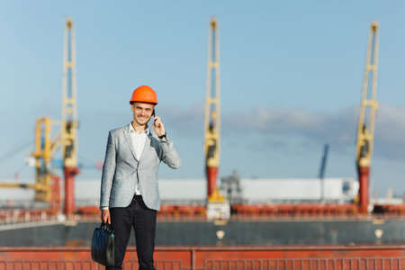 Handsome young unshaven successful business man in gray suit and protective construction orange helmet holding case, talking on mobile phone, walking in sea port against cargo ship, crane background.の写真素材