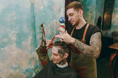 Man getting trendy haircut at barber shop. The male hairstylist in tattoos serving client.の写真素材