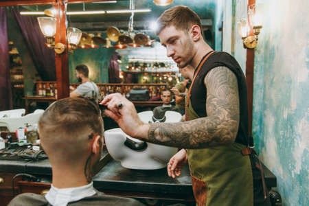 Close up shot of man getting trendy haircut at barber shop. The male hairstylist in tattoos serving client.の写真素材