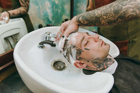 Close up shot of man getting trendy haircut at barber shop. The male hairstylist in tattoos serving client, washing headの写真素材