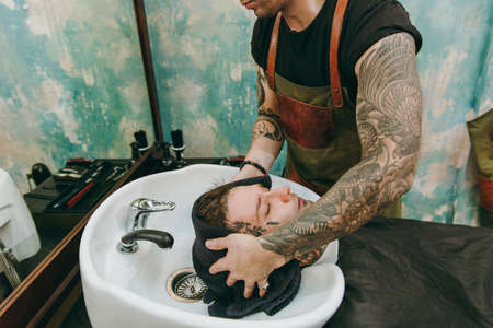 Close up shot of man getting trendy haircut at barber shop. The male hairstylist in tattoos serving...