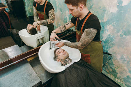 Man getting trendy haircut at barber shop. The male hairstylist in tattoos serving client, washing headの写真素材