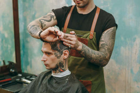 Close up shot of man getting trendy haircut at barber shop. The male hairstylist in tattoos serving client.の写真素材