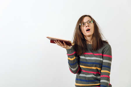 A beautiful European young brown-haired woman in glasses for sight dressed in casual dark grey longsleeve with colorful stripes, standing with book on a white background. Reading and studying conceptの写真素材