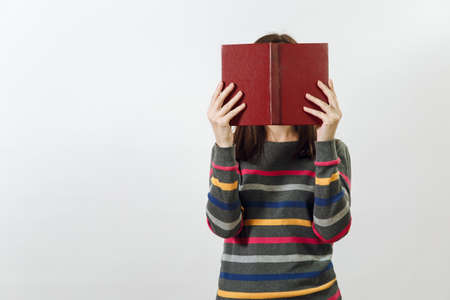 A beautiful European young brown-haired woman in glasses for sight dressed in casual dark grey longsleeve with colorful stripes, hiding behind book on a white background. Reading and studying conceptの写真素材