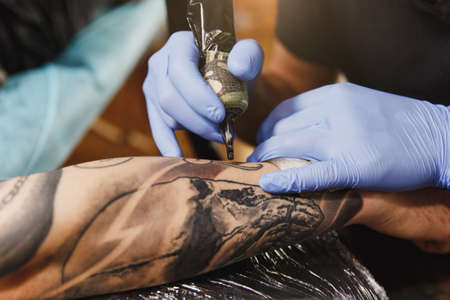 Close up of professional tattooer artist doing picture on hand of man by machine black ink from a jar. Tattoo art on body. Equipment for making tattoo art. Master makes tattooed in light studioの写真素材