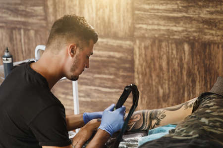Professional tattooer artist doing picture on hand of man by machine black ink from a jar. Tattoo...