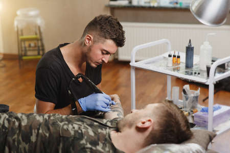 A professional tattooer artist doing picture on hand of man by machine black ink from a jar. Tattoo art on body. Equipment for making tattoo art. Master makes tattooed in light studio.の写真素材