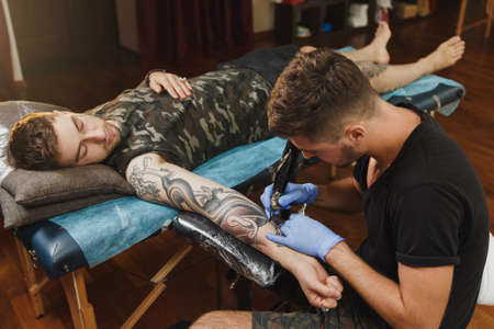 A professional tattooer artist doing picture on hand of man by machine black ink from a jar. Tattoo art on body. Equipment for making tattoo art. Master makes tattooed in light studio.の写真素材