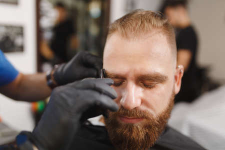 Male professional hairdresser serving client, shearing eyebrows with scissors. Ginger stylish young man with thick big beard and short hair getting trendy haircut in black cape. Light barber shopの写真素材