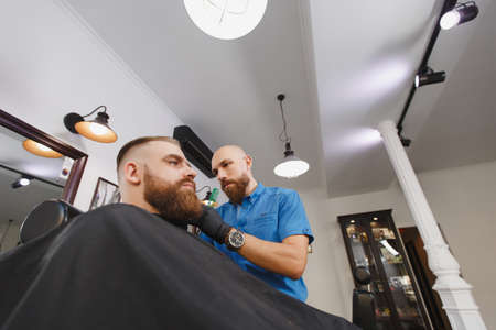 Male professional hairdresser serving client by clipper. Ginger handsome brutal stylish young man with thick big beard and short hair getting trendy haircut in black cape. Light white barber shopの写真素材