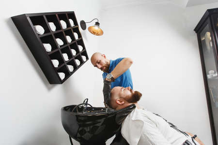 Male professional hairdresser serving client washing head in black trendy washbasin, wiping with towel. Ginger handsome stylish young man with thick big beard and short hair. Light white barber shopの写真素材